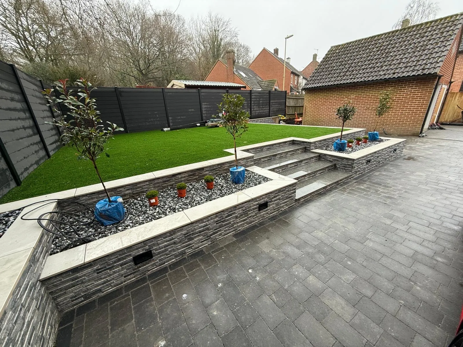 Charcoal block paving with stone-clad raised planters and steps up to an artificial lawn