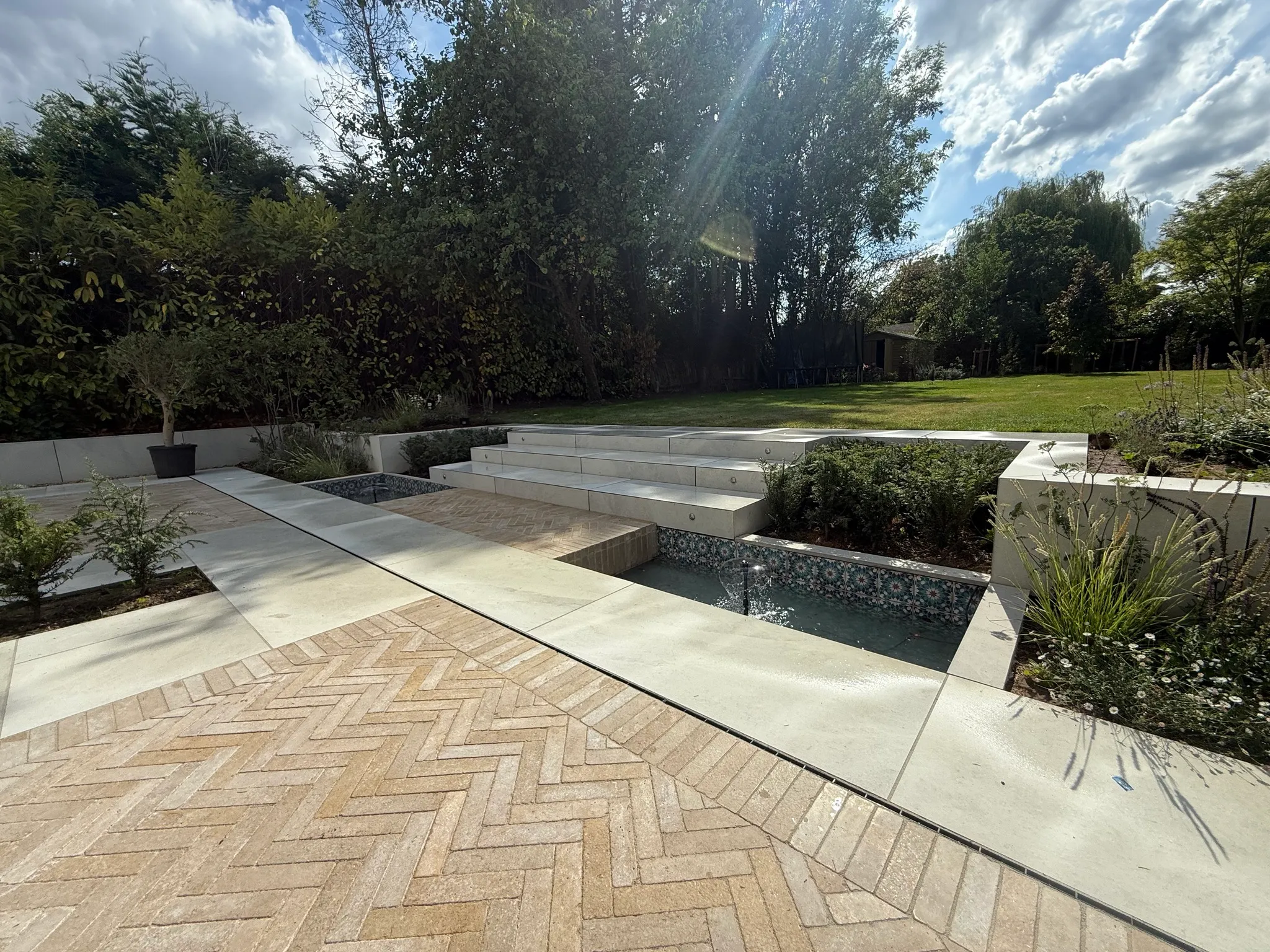 Herringbone brick and porcelain paving with a tiled water feature and steps up to a lawn